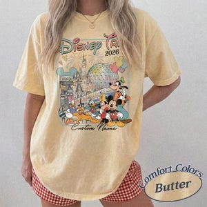 May include: Pale yellow t-shirt with a Disney-themed graphic featuring Mickey Mouse, Goofy, and Donald Duck, with the text "Disney Trip 2026" and "Custom Name". The shirt is a Comfort Colors brand.