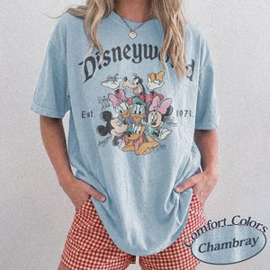 Może przedstawiać: Jasnoniebieska koszulka z grafiką postaci Disneya, w tym Myszki Miki, Myszki Minnie, Kaczora Donalda i Goofy'ego. Koszulka ma napis "Disneyworld Est. 1971". Wykonana z materiału Comfort Colors Chambray.