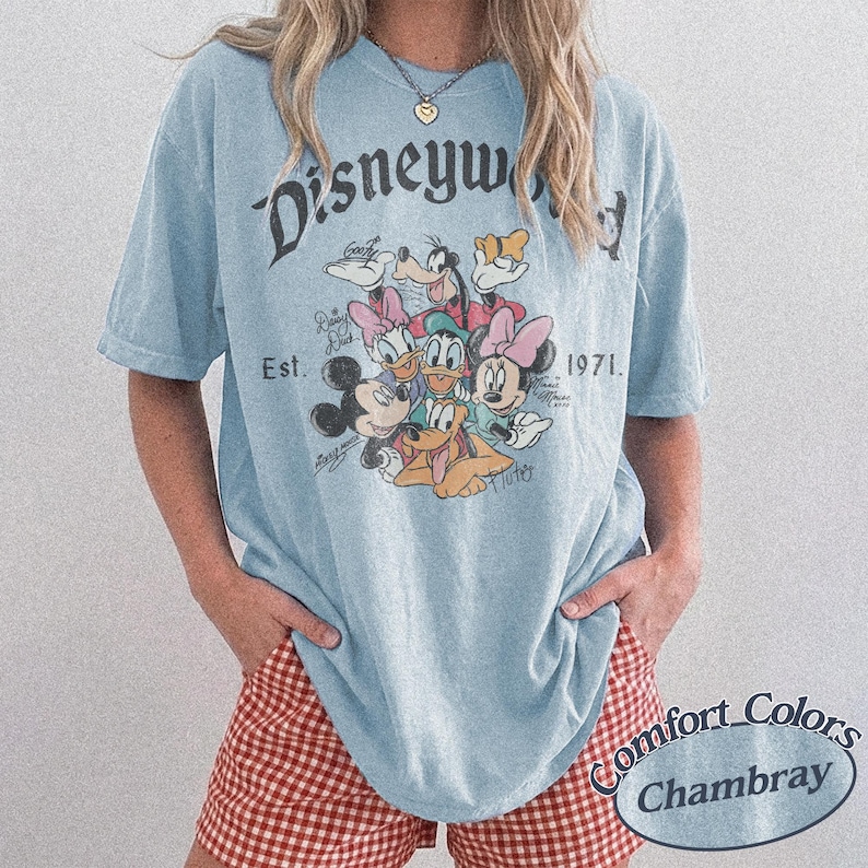 Może przedstawiać: Jasnoniebieska koszulka z grafiką postaci Disneya, w tym Myszki Miki, Myszki Minnie, Kaczora Donalda i Goofy'ego. Koszulka ma napis "Disneyworld Est. 1971". Wykonana z materiału Comfort Colors Chambray.