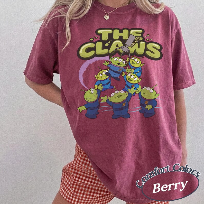 May include: Berry-colored t-shirt featuring a graphic of green aliens in blue uniforms with the text "THE CLAWS" in a lime green font. The shirt also has the text "Comfort Colors Berry" in a circular design.