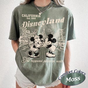 Puede incluir: Camiseta verde musgo con un dise&ntilde;o de Mickey y Minnie Mouse abraz&aacute;ndose frente a una escena de Disneyland. Incluye el texto "CALIFORNIA Est. 1955 Disneyland" y "The happiest place on earth".