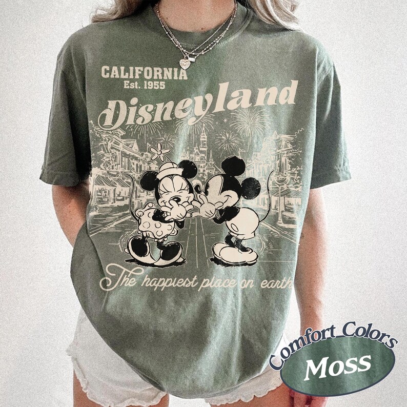 Puede incluir: Camiseta verde musgo con un dise&ntilde;o de Mickey y Minnie Mouse abraz&aacute;ndose frente a una escena de Disneyland. Incluye el texto "CALIFORNIA Est. 1955 Disneyland" y "The happiest place on earth".