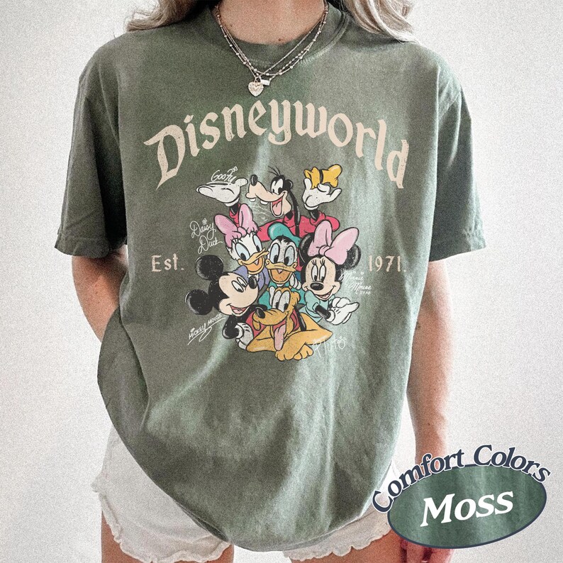 Może przedstawiać: Zielona koszulka w kolorze mchu z napisem "Disneyworld" w stylu vintage i grafiką postaci Disneya. Koszulka zawiera tekst "Est. 1971" oraz słowa "Comfort Colors Moss" w owalu.