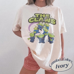 May include: Ivory-colored t-shirt featuring a graphic of the aliens from the movie Toy Story. The design includes the text "THE CLAWS" in green and a purple ring. The shirt is labeled "Comfort Colors Ivory".