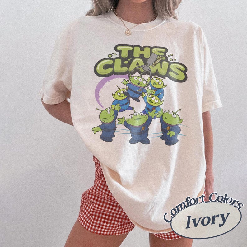 May include: Ivory-colored t-shirt featuring a graphic of the aliens from the movie Toy Story. The design includes the text "THE CLAWS" in green and a purple ring. The shirt is labeled "Comfort Colors Ivory".