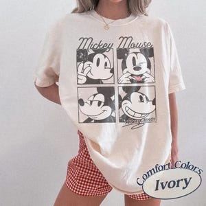 Op de afbeelding: Ivoor kleurig T-shirt met een vierpaneels afbeelding van Mickey Mouse met verschillende uitdrukkingen. De woorden "Mickey Mouse" staan boven de afbeeldingen. Het shirt is gelabeld met "Comfort Colors Ivory".