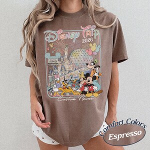 May include: Espresso-coloured t-shirt with a vibrant graphic of Disney characters, including Mickey Mouse, Donald Duck, and Goofy, with the text "Disney Trip 2026" and "Custom Name". The shirt is from Comfort Colors.