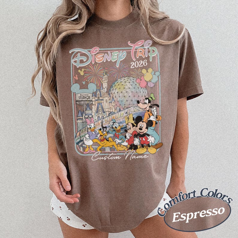 May include: Espresso-coloured t-shirt with a vibrant graphic of Disney characters, including Mickey Mouse, Donald Duck, and Goofy, with the text "Disney Trip 2026" and "Custom Name". The shirt is from Comfort Colors.