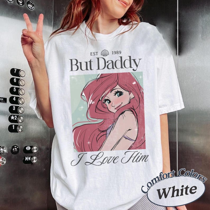 K&ouml;nnte beinhalten: Wei&szlig;es T-Shirt mit einer Cartoon-Illustration einer Figur mit rosa Haaren und dem Text "But Daddy I Love Him". Das Shirt hat auch den Text "EST 1989" und "Comfort Colors White".
