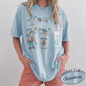 Puede incluir: Camiseta azul claro con ilustraciones de personajes de dibujos animados y texto. La camiseta incluye im&aacute;genes de Woody, una patata y otros personajes. El texto incluye "Andy's Room Keep Out" y "Wanted Reward". La camiseta est&aacute; etiquetada como "Comfort Colors Chambray".