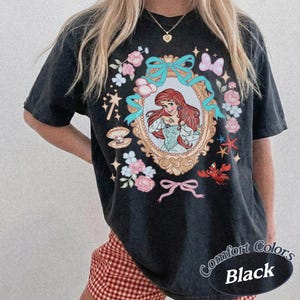 Puede incluir: Camiseta negra con una ilustraci&oacute;n enmarcada de una mujer pelirroja con un vestido azul, rodeada de flores, lazos y elementos marinos. El texto "Comfort Colors Black" est&aacute; impreso en la parte inferior derecha.