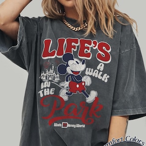 May include: Dark gray t-shirt featuring Mickey Mouse and the text "LIFE'S A WALK IN THE PARK" in red and white. The shirt also includes the Walt Disney World logo and is labeled "Comfort Colors Pepper".
