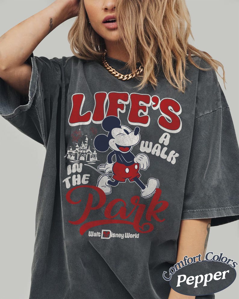 May include: Dark gray t-shirt featuring Mickey Mouse and the text "LIFE'S A WALK IN THE PARK" in red and white. The shirt also includes the Walt Disney World logo and is labeled "Comfort Colors Pepper".