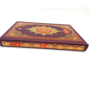 May include: A maroon book with gold and red decorative accents. The cover features an ornate gold medallion with Arabic script. The spine has red text and gold embellishments. The book is closed and resting on a white surface.