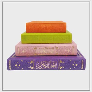 May include: Four brightly coloured books stacked on top of each other. The top book is orange, followed by green, pink, and purple at the bottom. The books have gold embossed text and designs. The bottom book has Arabic text.