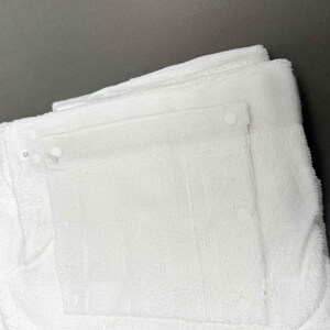 Puede incluir: Primer plano de toallas de rizo blancas dobladas con un bolsillo de pl&aacute;stico transparente asegurado con broches de pl&aacute;stico blanco. Las toallas est&aacute;n apiladas, con el bolsillo visible en la capa superior. El fondo es gris liso.