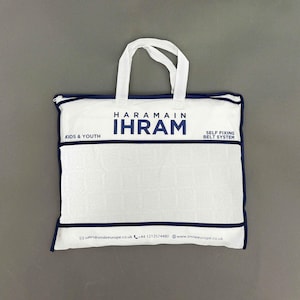 Puede incluir: Bolsa blanca con ribete y asas azul marino. La bolsa muestra el texto "HARAMAIN IHRAM" en azul marino, con "KIDS & YOUTH" y "SELF FIXING BELT SYSTEM" impresos. La bolsa est&aacute; dise&ntilde;ada para ni&ntilde;os y j&oacute;venes.