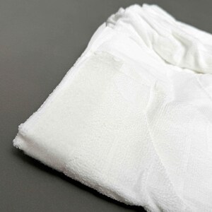 Puede incluir: Primer plano de una toalla de rizo blanca doblada. La toalla tiene una superficie texturizada y un borde doblado. El fondo es de color gris s&oacute;lido. La toalla parece suave y absorbente.