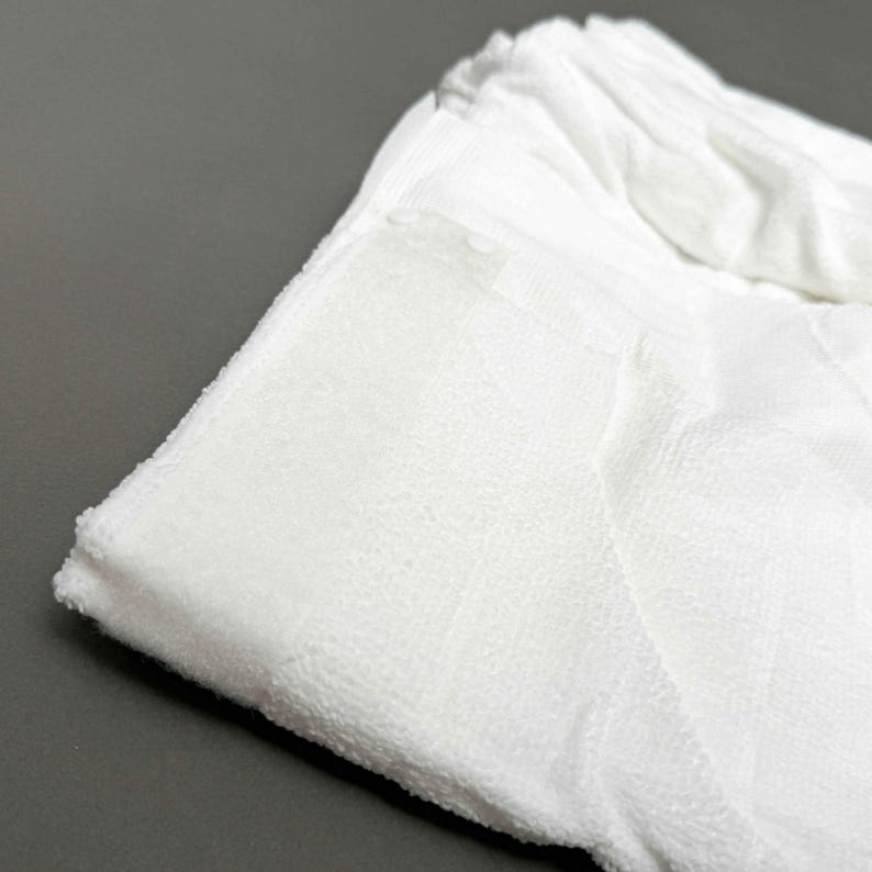 Puede incluir: Primer plano de una toalla de rizo blanca doblada. La toalla tiene una superficie texturizada y un borde doblado. El fondo es de color gris s&oacute;lido. La toalla parece suave y absorbente.