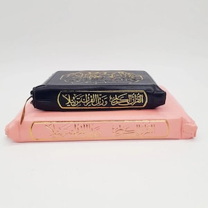 Op de afbeelding: Twee boeken, één zwart en één roze, op elkaar gestapeld. Het zwarte boek heeft gouden letters en een ritssluiting. Het roze boek heeft ook gouden letters. Beide lijken religieuze teksten te zijn.