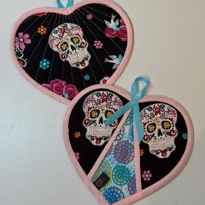 May include: Two heart-shaped oven mitts with a Day of the Dead theme. The black fabric features a sugar skull design, flowers, and birds. The edges are trimmed in pink, and each has a blue ribbon loop.