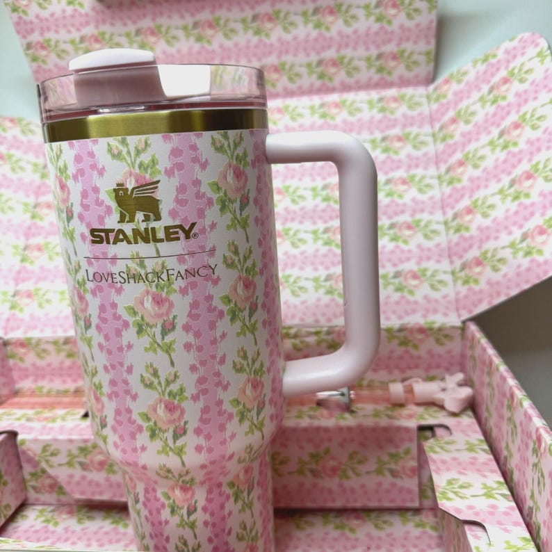 May include: A pink and white floral Stanley tumbler with a handle. The tumbler has a gold band and a clear lid. The Stanley logo and "Loveshackfancy" are printed on the front. The tumbler is in a matching floral box.