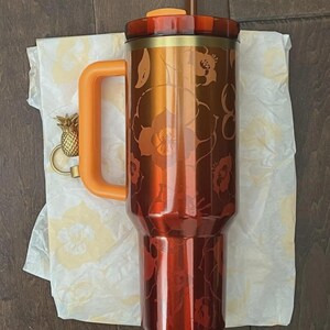 May include: A large, insulated tumbler with a floral design in shades of orange and red. It has an orange handle and lid, a brown straw, and a gold pineapple charm. The tumbler is approximately 10 inches tall.