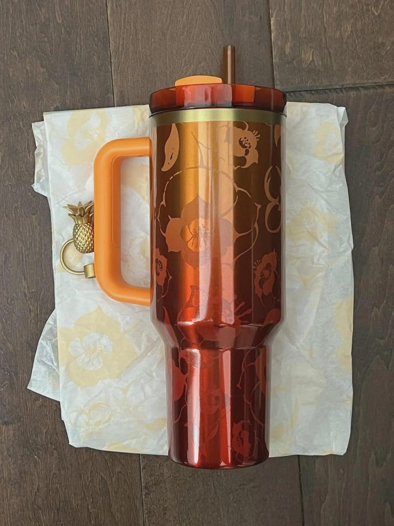 May include: A large, insulated tumbler with a floral design in shades of orange and red. It has an orange handle and lid, a brown straw, and a gold pineapple charm. The tumbler is approximately 10 inches tall.