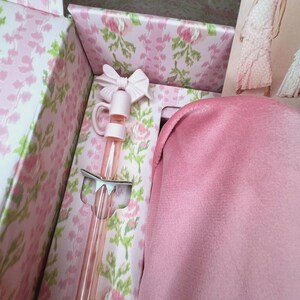May include: A pink gift box with a floral pattern, containing a pink bow-topped bottle and a pink fabric item. The box's interior is also decorated with a floral design. A fashion illustration is visible in the background.