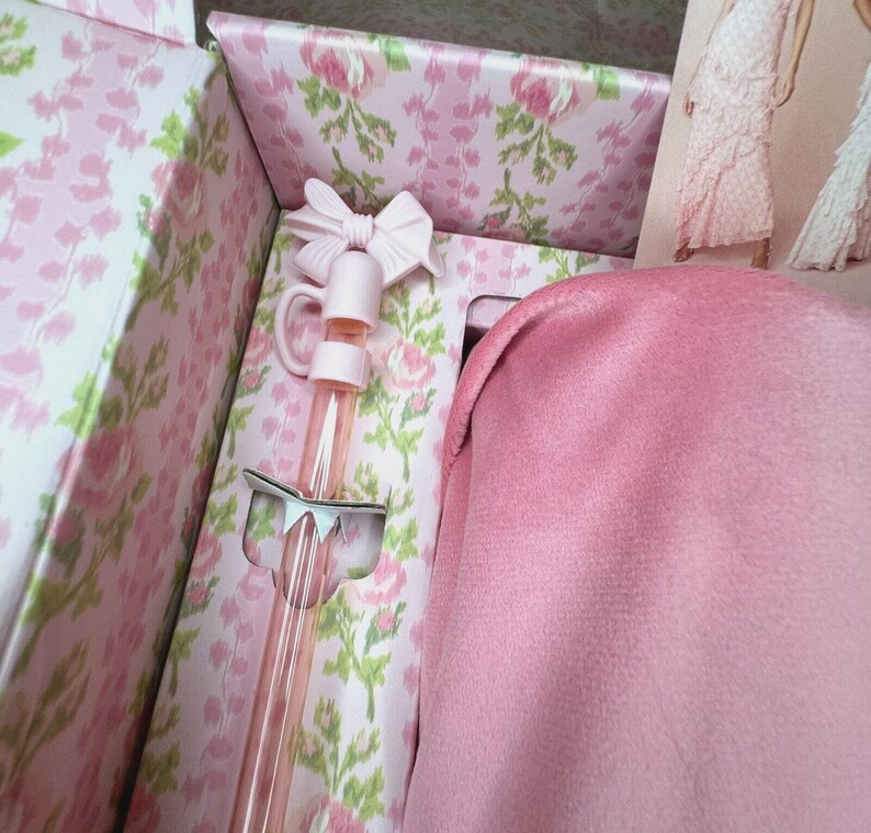 May include: A pink gift box with a floral pattern, containing a pink bow-topped bottle and a pink fabric item. The box's interior is also decorated with a floral design. A fashion illustration is visible in the background.