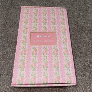 May include: A pink rectangular box with a floral pattern of pink roses and green leaves. The box has a pink label with the text "STANLEY" and "LoveShackFancy" in gold lettering.