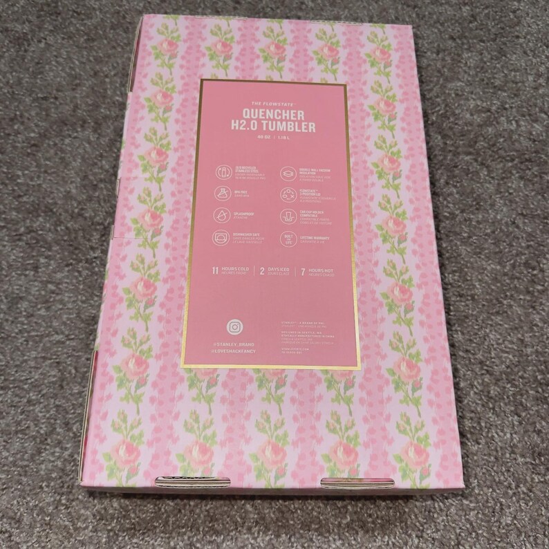May include: A pink box with a floral pattern and the text "QUENCHER H2.0 TUMBLER". The box has a gold border and contains information about the product. The floral pattern includes pink roses and green leaves.