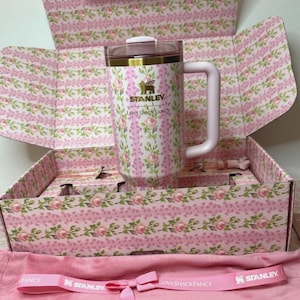 May include: A pink floral Stanley travel mug with a handle, inside a matching floral box. The box and mug feature a pink and white floral pattern. The mug has the Stanley and LoveShackFancy logos. A pink bag and ribbon with the Stanley and LoveShackFancy logos are also visible.