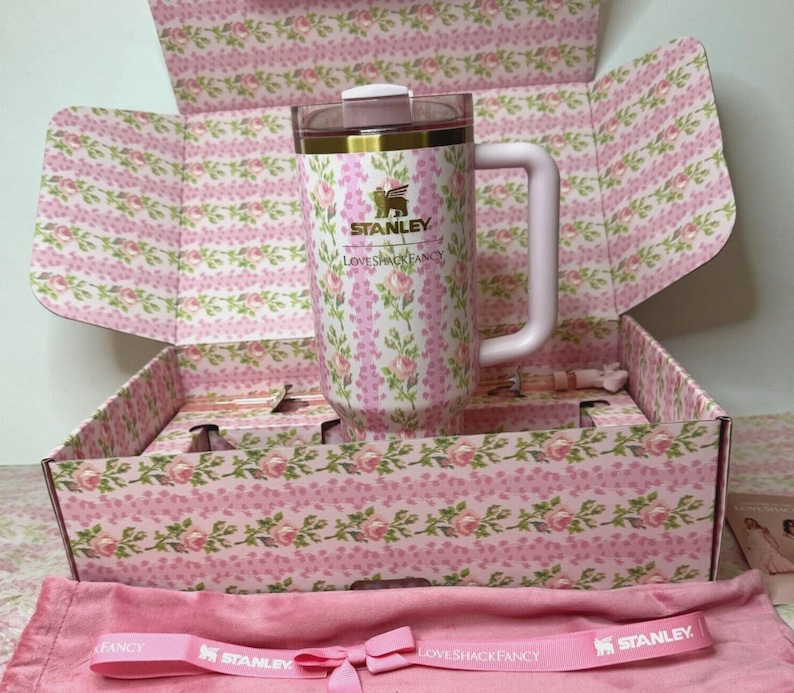 May include: A pink floral Stanley travel mug with a handle, inside a matching floral box. The box and mug feature a pink and white floral pattern. The mug has the Stanley and LoveShackFancy logos. A pink bag and ribbon with the Stanley and LoveShackFancy logos are also visible.