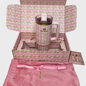 May include: A Stanley travel mug with a white handle and a floral pattern, presented in a matching box. The mug is approximately 8 inches tall. The box and accessories are pink. The set includes a pink drawstring bag and a pink ribbon.