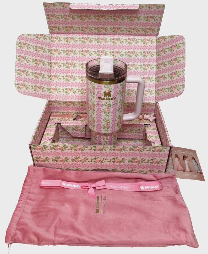 May include: A Stanley travel mug with a white handle and a floral pattern, presented in a matching box. The mug is approximately 8 inches tall. The box and accessories are pink. The set includes a pink drawstring bag and a pink ribbon.