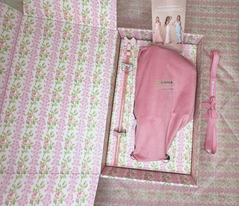 May include: Open gift box with a floral pattern in pink and white. Inside, a pink Stanley brand bag, a pink stirrer, and a pink strap are visible. A card with three women in dresses is also inside the box.