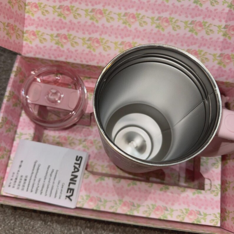 May include: A pink Stanley travel mug with a matching lid and handle, presented in a floral-patterned box. The stainless steel interior is visible, along with a product information card. The mug is designed for beverages.