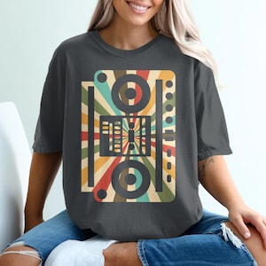 以下が含まれることがあります： レトロスタイルのサウンドミキサーのグラフィックが特徴の濃いグレーのTシャツ。茶色、オレンジ、緑、青の色合いで、円、長方形、線が描かれたカラフルなサンバーストのデザインです。カジュアルウェアアイテムです。