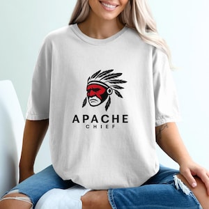 May include: White t-shirt featuring a graphic of an Apache Chief. The design includes a red face, black feathers, and the words "APACHE CHIEF" in a bold, black font. The shirt is a casual, unisex style.