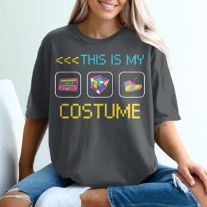 Op de afbeelding: Donkergrijs T-shirt met de tekst "THIS IS MY COSTUME" in pixellettertype. Het shirt heeft drie retro-iconen: een cassettebandje, een Rubik's Cube en een rolschaats. Het shirt wordt door een persoon gedragen.