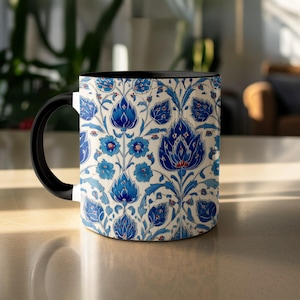 Op de afbeelding: Een keramische mok met een zwarte handgreep en binnenkant. De buitenkant heeft een gedetailleerd bloemmotief in blauw- en wittinten. Het ontwerp bevat gestileerde bloemen en bladeren, wat een decoratieve en elegante uitstraling creëert.