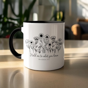 May include: White ceramic mug with a black handle and interior. The mug features a black line drawing of various flowers and the text "Hold on to what you have" with the reference "Revelation 3:11".
