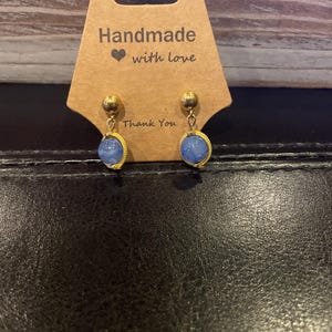 May include: Gold-colored stud earrings with oval, light blue gemstones. The earrings are displayed on a brown card with the text "Handmade with love" and "Thank You".