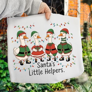 May include: White fabric featuring four cartoon geese dressed as Santa's helpers, wearing green and red elf outfits and hats. The geese are decorated with Christmas lights and the text "Santa's Little Helpers."