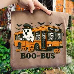 Op de afbeelding: Een bruine Halloween-draagtas met een oranje bus met het opschrift "BOO-BUS", bestuurd door een spook. Skeletten en geesten zijn passagiers. Pompoenen en vleermuizen versieren het ontwerp. De tas wordt door een hand vastgehouden.