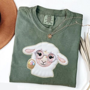May include: An olive green Comfort Colors t-shirt with a white sheep design, pink details, glasses, and an Easter egg basket. The sheep is edged with pearls. A gold necklace is draped across the shirt.
