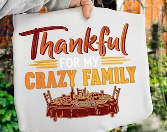 May include: White tote bag with the words "Thankful for my Crazy Family" in orange and brown script. Below the text is an illustration of a Thanksgiving dinner table set with food and chairs. The bag is held by a person.