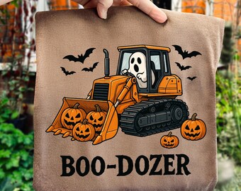 May include: A brown Halloween-themed pillow featuring an orange bulldozer with a ghost driver, pumpkins, and bats. The text "BOO-DOZER" is printed in black. The pillow is held by a hand.