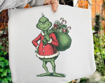 Könnte beinhalten: Eine weiße Tragetasche mit einer Illustration des grünen Grinch, der einen roten Weihnachtsmannanzug trägt und einen Sack voller Geschenke und Zuckerstangen trägt. Ein festliches Design für die Weihnachtszeit.